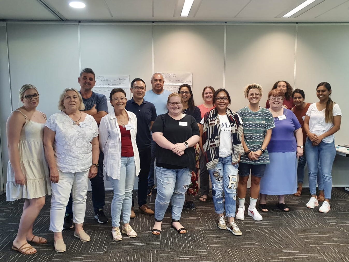 A group of 15 people (men and women), dressed in casual clothes, and smiling at the camera after completing the Mental Health First Aid course with Mental Strides