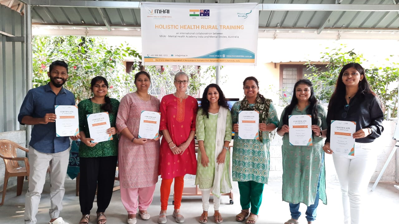 Eight adults stand in a row, smiling. Six hold certificates of completion in Holistic Health, a course presented by Mental Strides and the Mental Health Academy India. 
