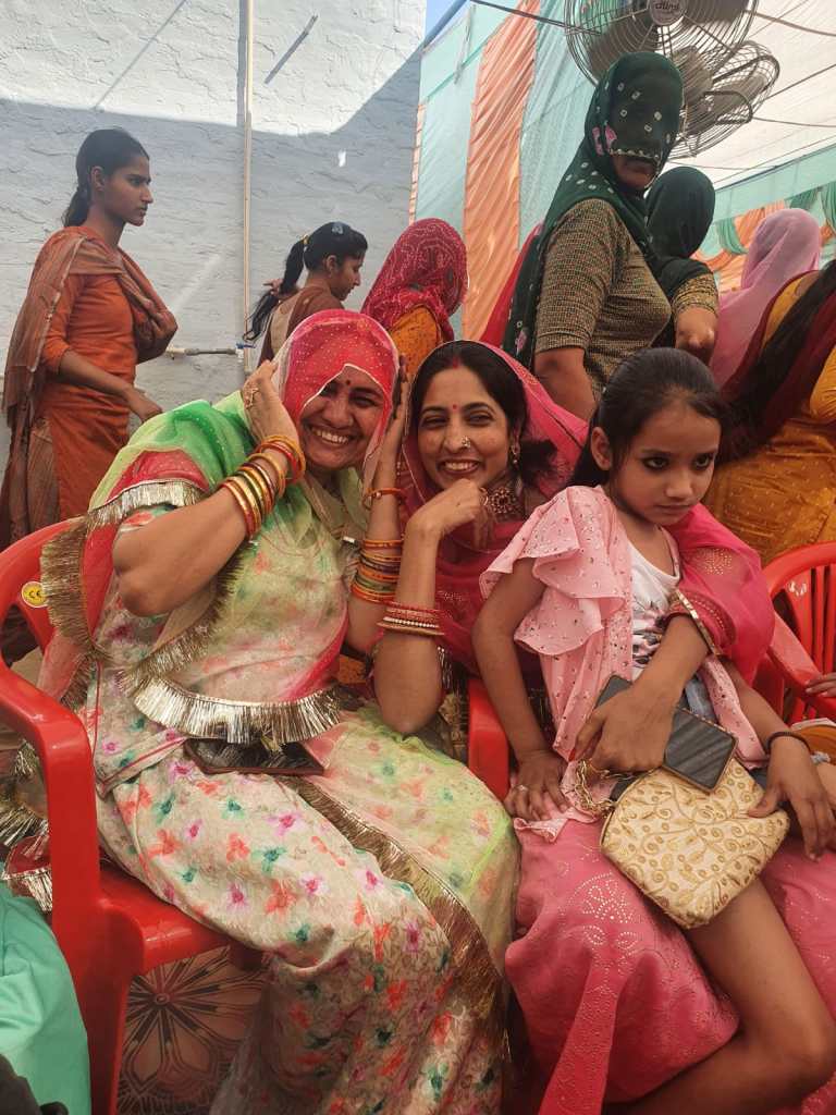 Two good friends, dressed in saris, laugh together while posing for a photo as part of wedding celebrations. A young girl sits on one of the women's knees.