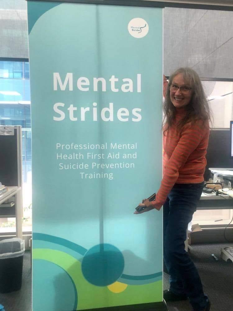 Mental Strides Mental Health Consultant, Engel Prendergast poses with a teal blue pull-up batter with the words "Mental Strides" written on it.