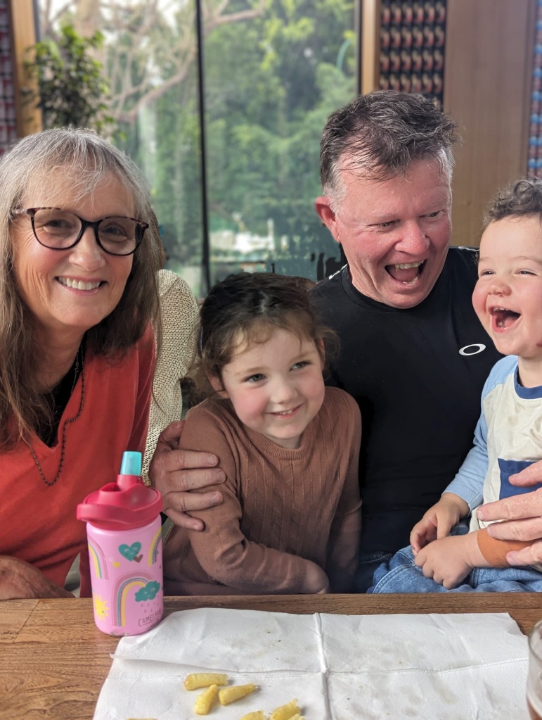 Two grandparents and their pre-school and toddler grandchildren laugh together while sitting at a table.